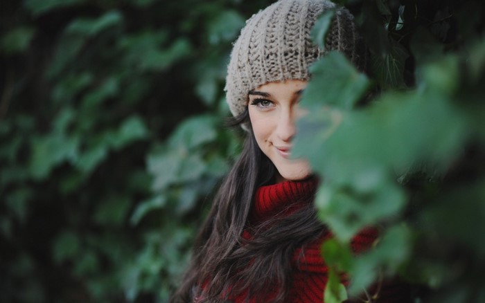 Woman in a knitted hat and red sweater peeking through leaves
