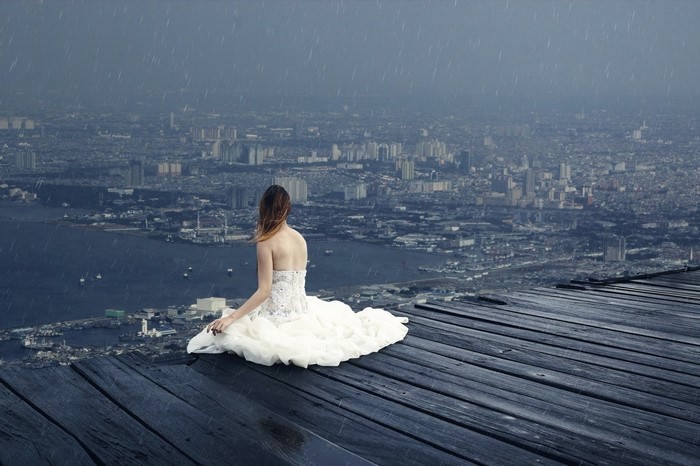Woman in white dress sitting on wooden planks overlooking city in rain
