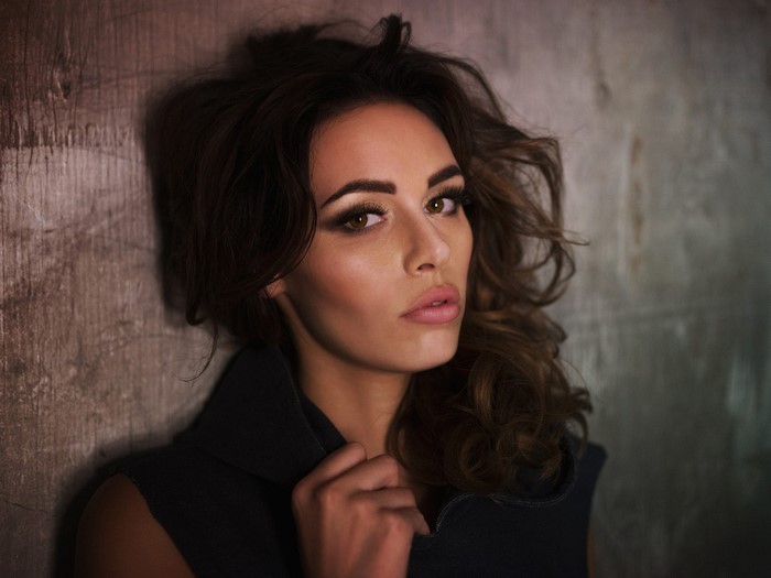 Woman with long brown hair posing against a wall