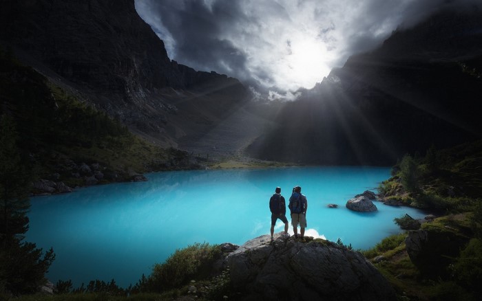 Two hikers admire a turquoise mountain lake with sun rays