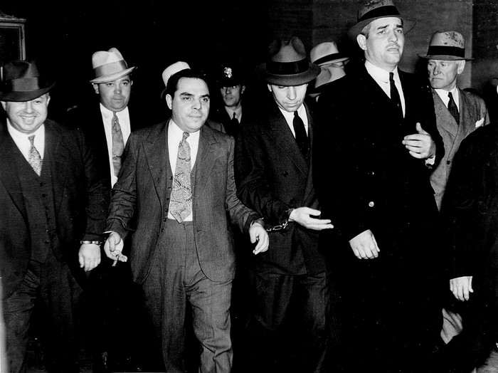 Men in suits and hats, some handcuffed, being escorted