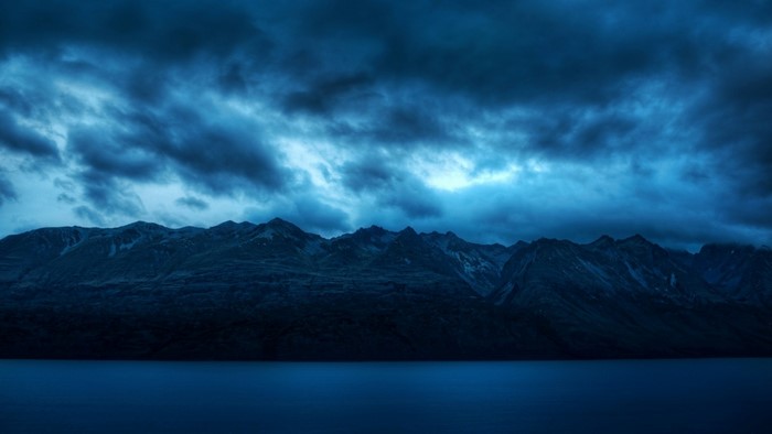 Dark mountains over a calm lake under stormy blue clouds