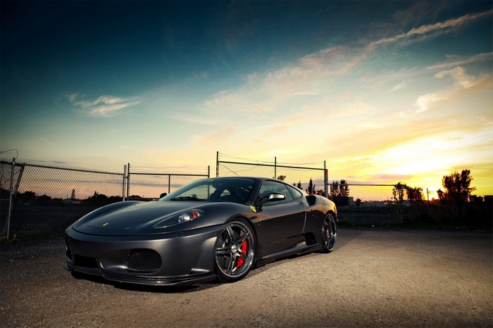 Dark gray Ferrari F430 parked near a fence at sunset