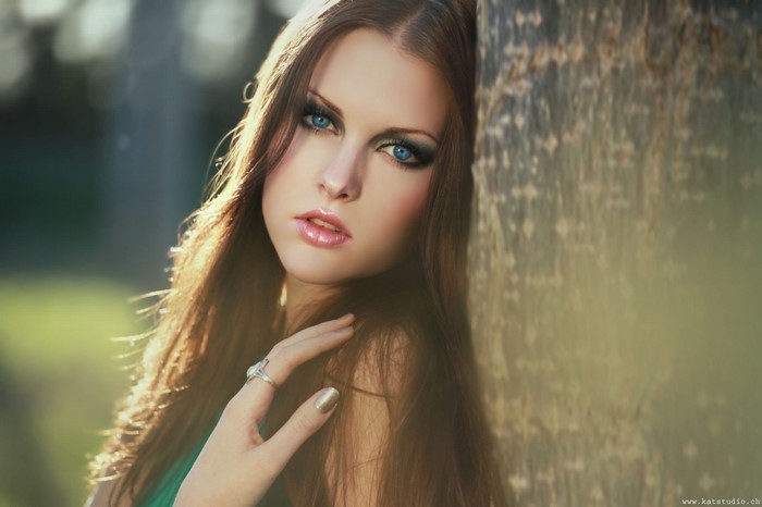 Woman with long brown hair and blue eyes leaning against a tree