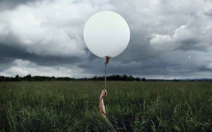 Hand holding a white balloon in a grassy field under stormy clouds