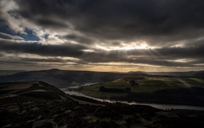 Sun rays breaking through dark clouds over a valley with a river and hills