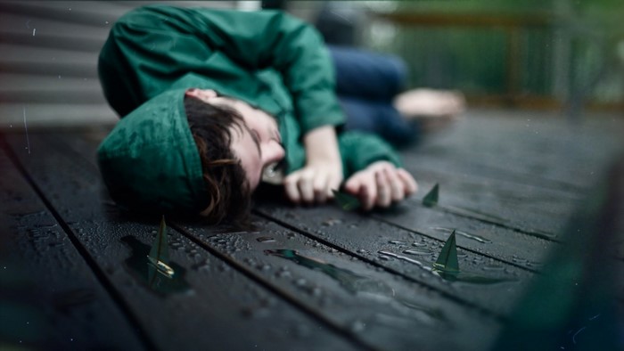 Person in green jacket lying on wet wooden surface with paper boats