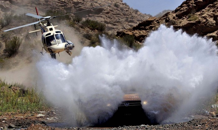 Helicopter flies over a car splashing through water in a rocky canyon