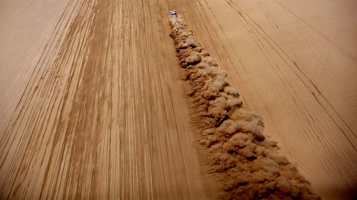 Off-road vehicle racing through sand dunes, creating dust clouds