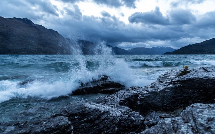 Waves crashing on rocks with mountains under cloudy sky