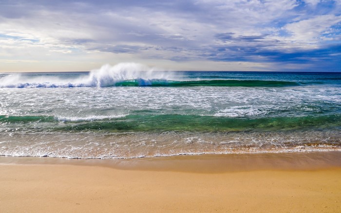 Ocean wave crashing on a sandy beach under a cloudy sky