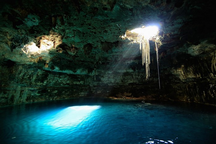 Sunlight beams into a cave with a turquoise pool and stalactites