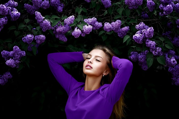 Woman in purple dress with hands on head near lilacs