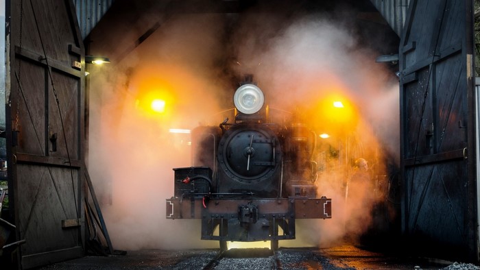 Steam locomotive emerging from a dark shed with steam and lights