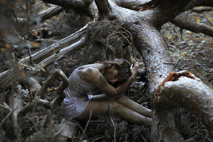 Woman covered in mud sitting amongst tree roots