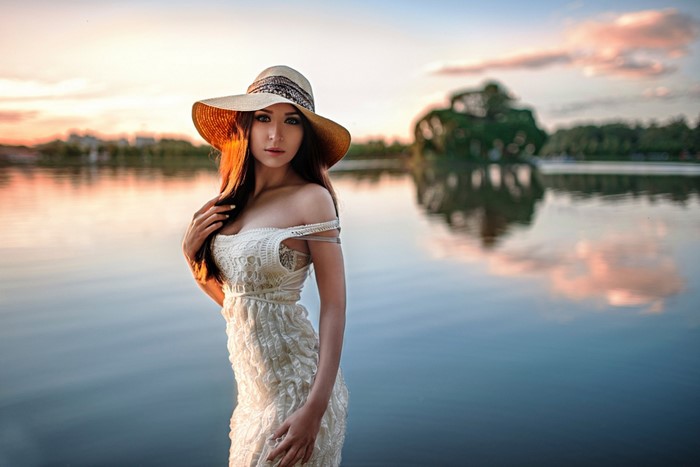 Woman in a white dress and hat by the water at sunset