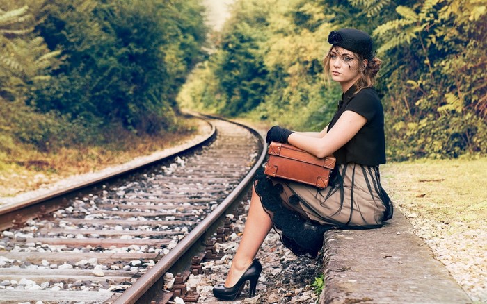 Woman in vintage clothing sitting by train tracks with suitcase