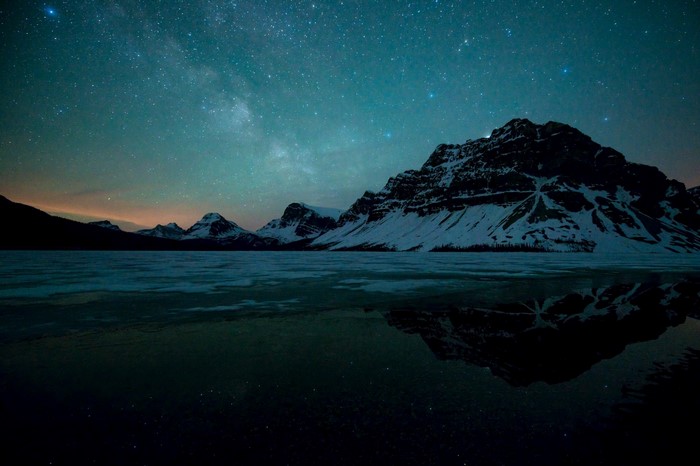 Snowy mountains under a starry night sky reflected in a frozen lake