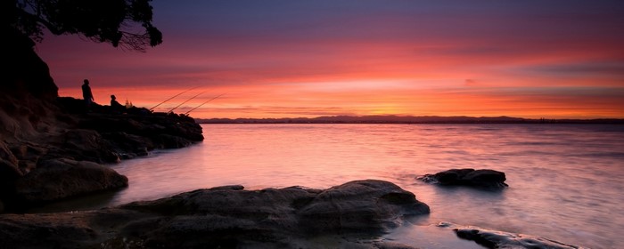 Two people fishing on a rocky shore at sunset