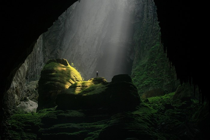 Person standing on mossy rocks in a large cave with sun rays