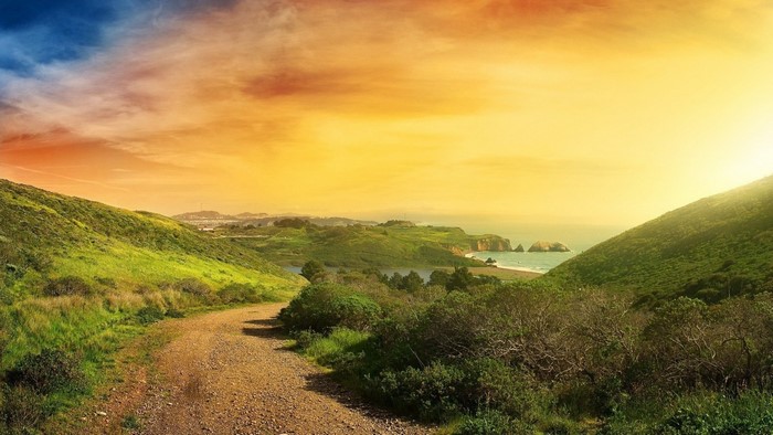 Dirt path through green hills towards the ocean at sunset