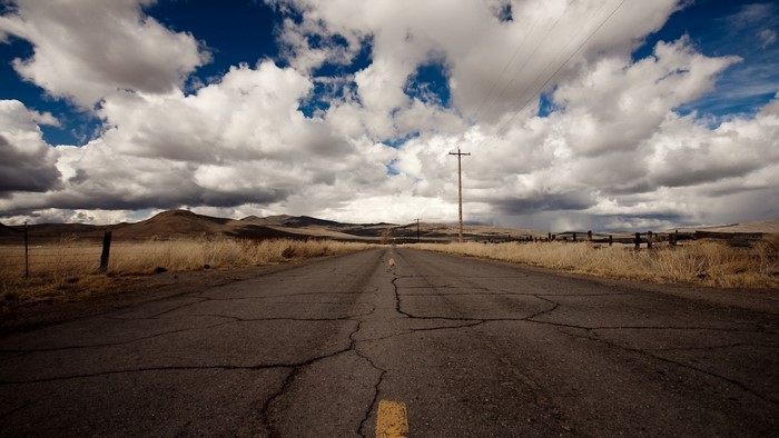 Cracked asphalt road through a dry, grassy landscape under a cloudy sky