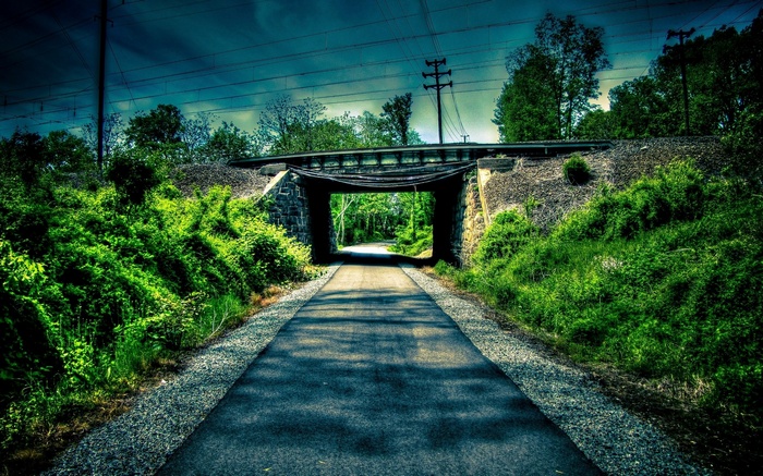 A paved path goes under a stone bridge with trees