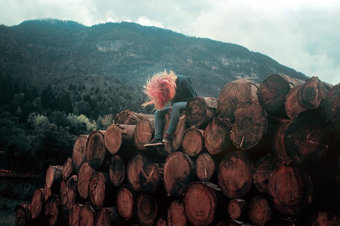 Person with pink hair sitting on logs in front of a forest