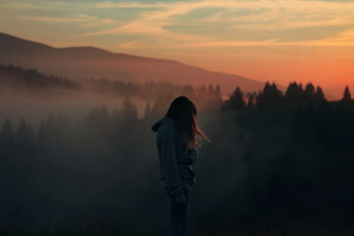 Girl in hoodie looking at misty mountains at sunset