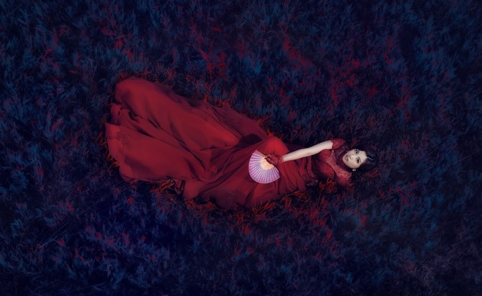 Woman in red dress lying on dark grass holding a fan