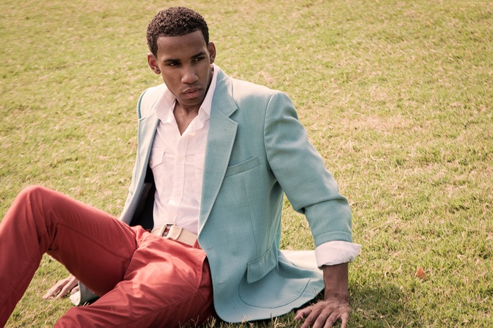 Male model wearing a teal blazer and red pants sitting on grass