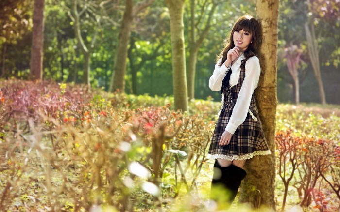 Young Asian woman in plaid dress leaning against tree in park