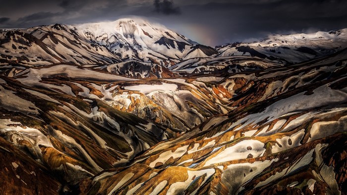 Snow-dusted mountains with a rainbow under dark clouds