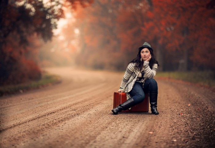 Woman sitting on a suitcase on a dirt road in autumn