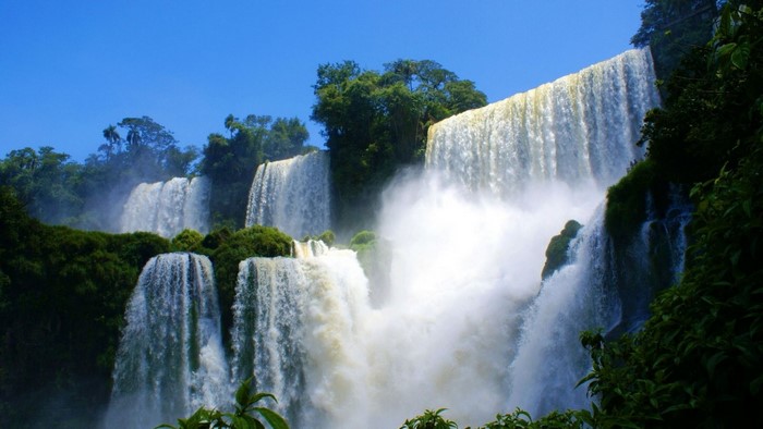 Multiple waterfalls cascading down a lush green cliffside under a clear blue sky