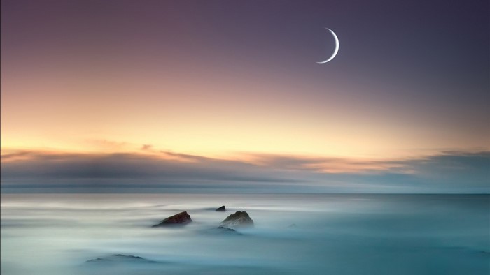 Crescent moon over the ocean at sunrise with rocks