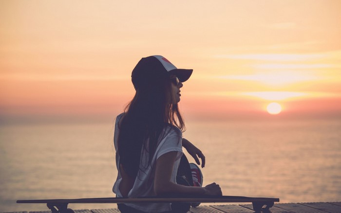 Woman in cap sitting on longboard at sunset by the sea
