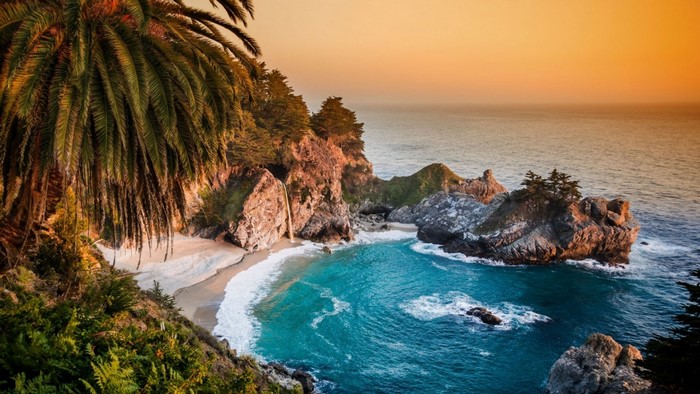 Tropical beach cove with waterfall and palm trees at sunset