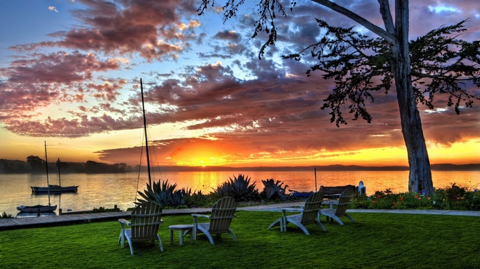 Sunset over a calm lake with sailboats and Adirondack chairs
