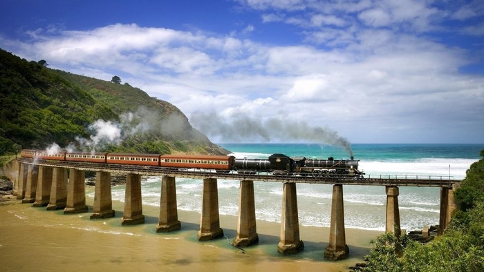 Steam train on a bridge over water with ocean and hills