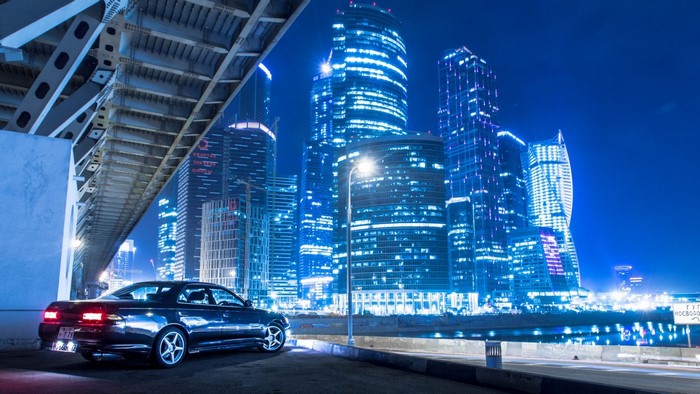 Dark Toyota Mark II parked under a bridge with Moscow skyscrapers at night