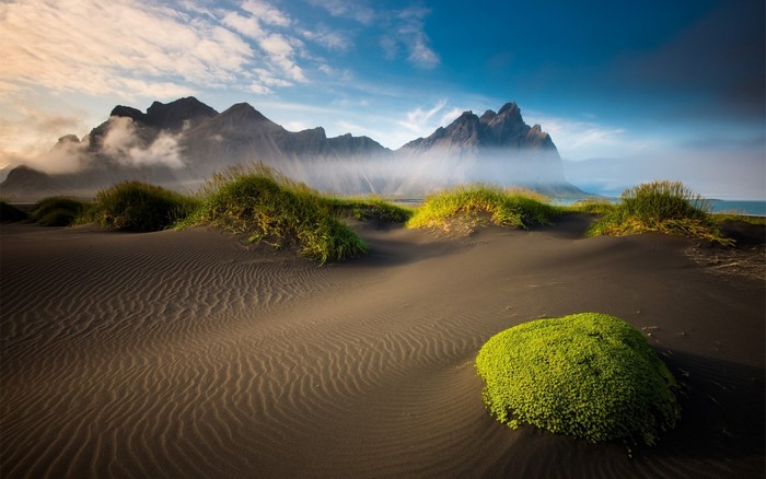 Black sand dunes with grass and mountains under a cloudy sky
