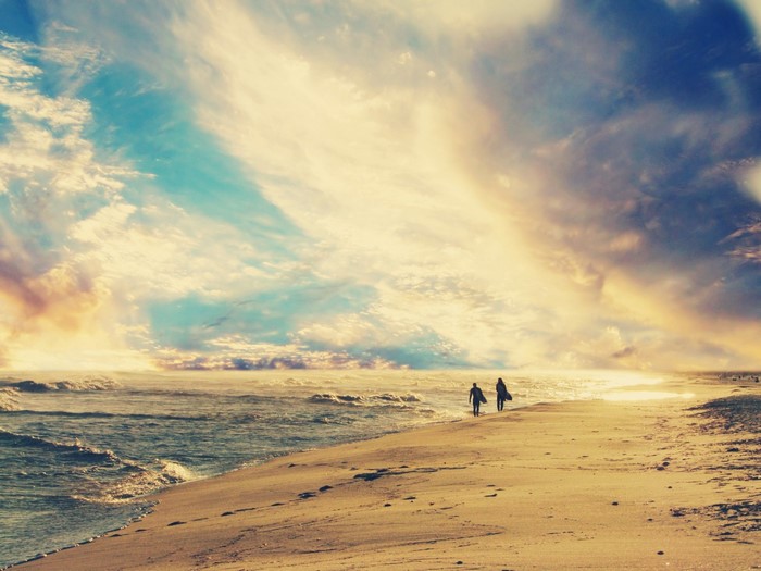 Two surfers walk along a sandy beach at sunset