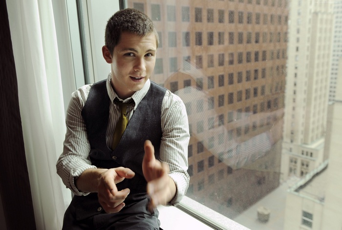 Logan Lerman sitting by a window with buildings in the background