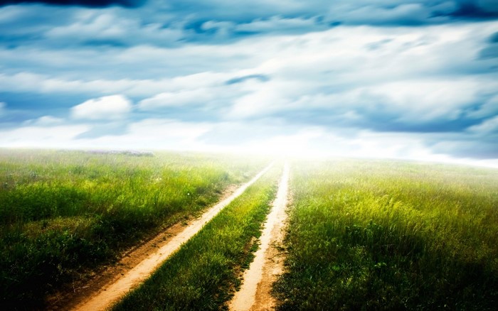 Dirt road through a green field under a cloudy sky