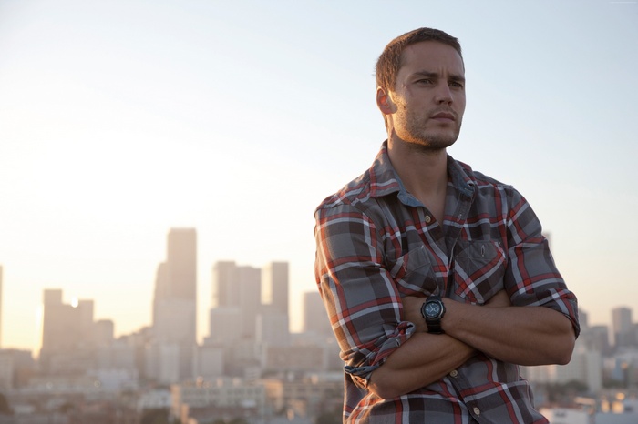 Taylor Kitsch standing with arms crossed on a rooftop