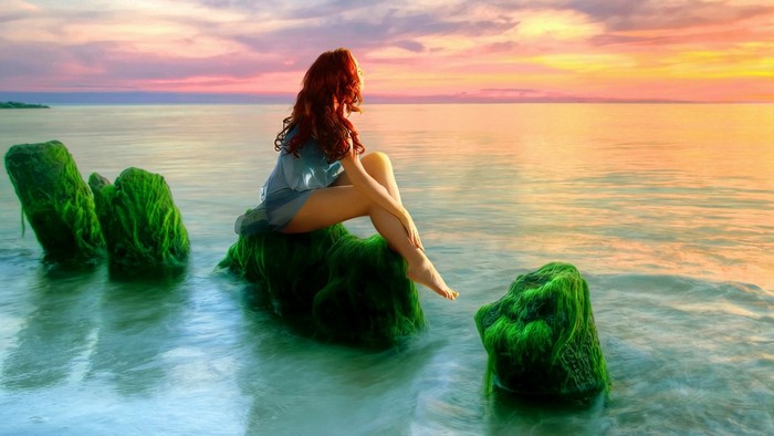 Woman with red hair sitting on mossy rocks in the sea at sunset