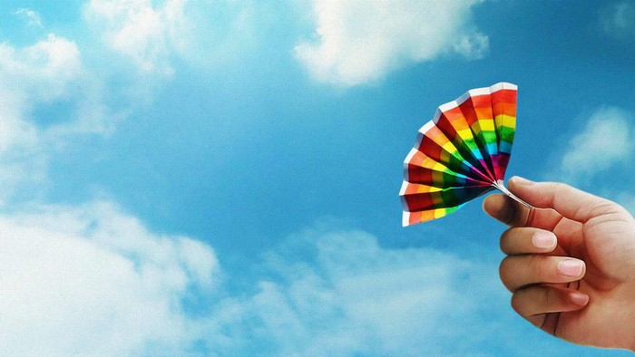 Hand holding a rainbow fan against a blue sky