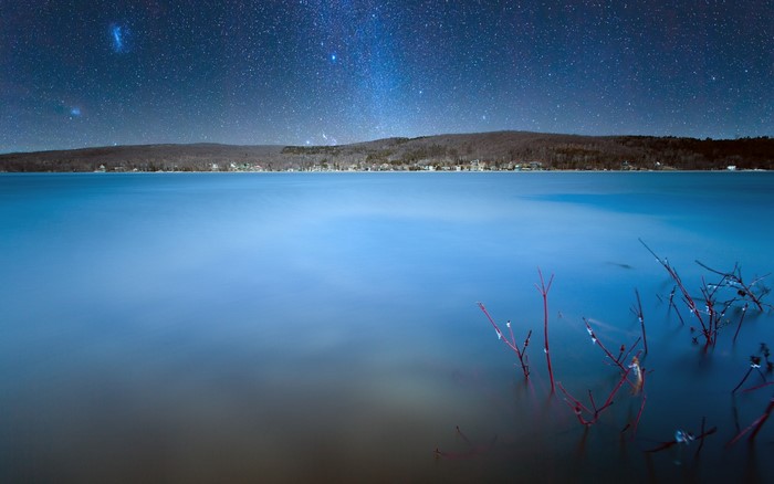 Night sky over a calm lake with trees on the horizon