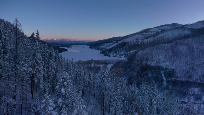Snow-covered forest overlooking a frozen lake and dam at dawn